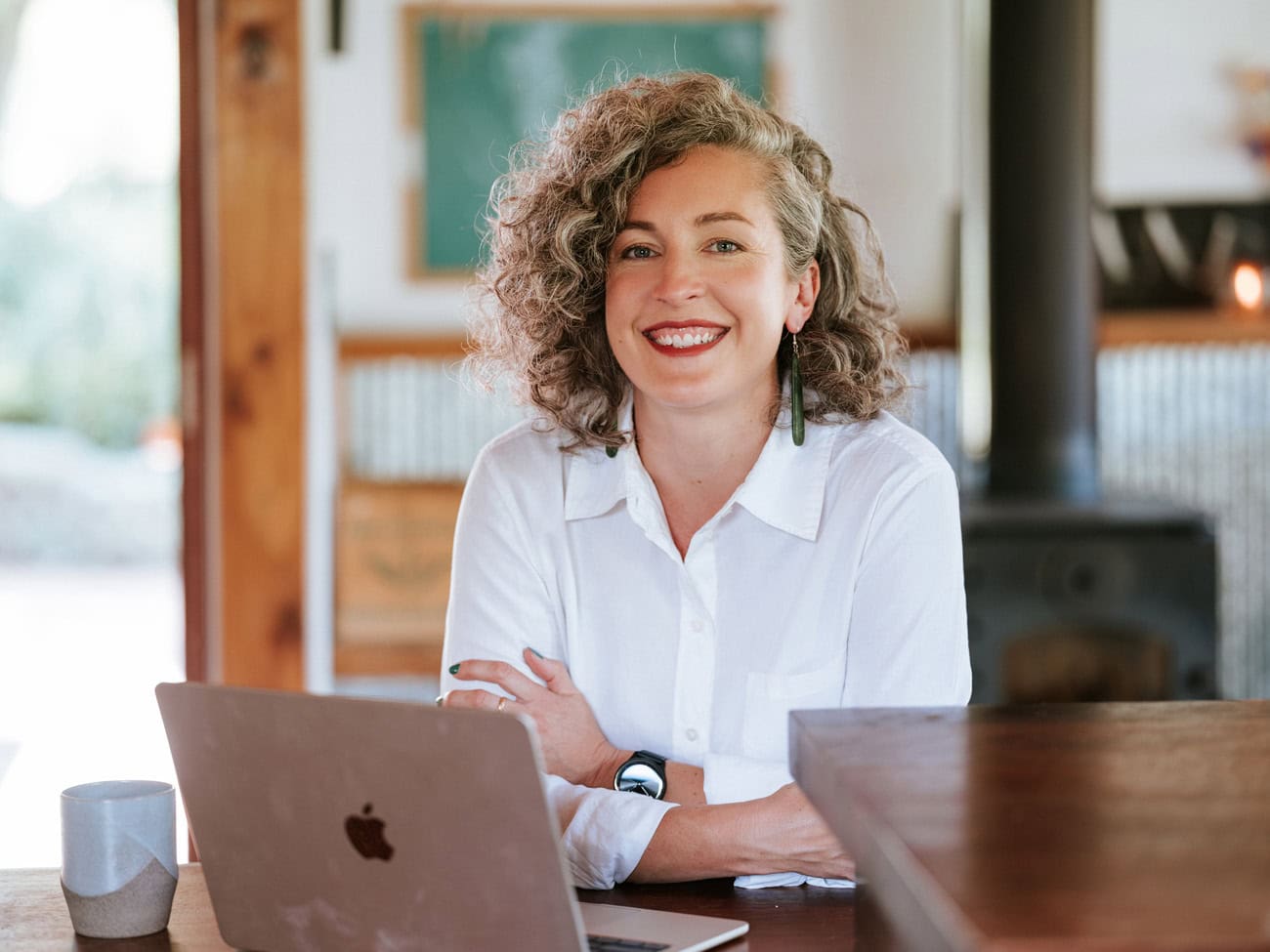 Photo by Florence Charvin Photography of A woman with curly hair and a white shirt sits at a table with a laptop and a mug, smiling at the camera in a cosy indoor setting.