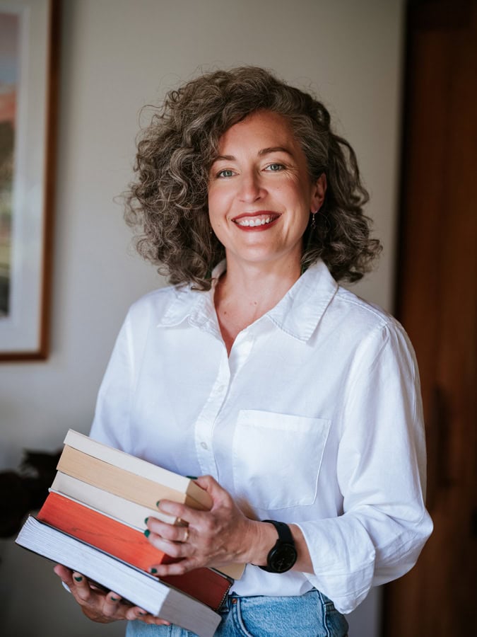 Photo by Florence Charvin Photography of A woman with curly hair, wearing a white shirt and jeans, smiles whilst holding a stack of books indoors.