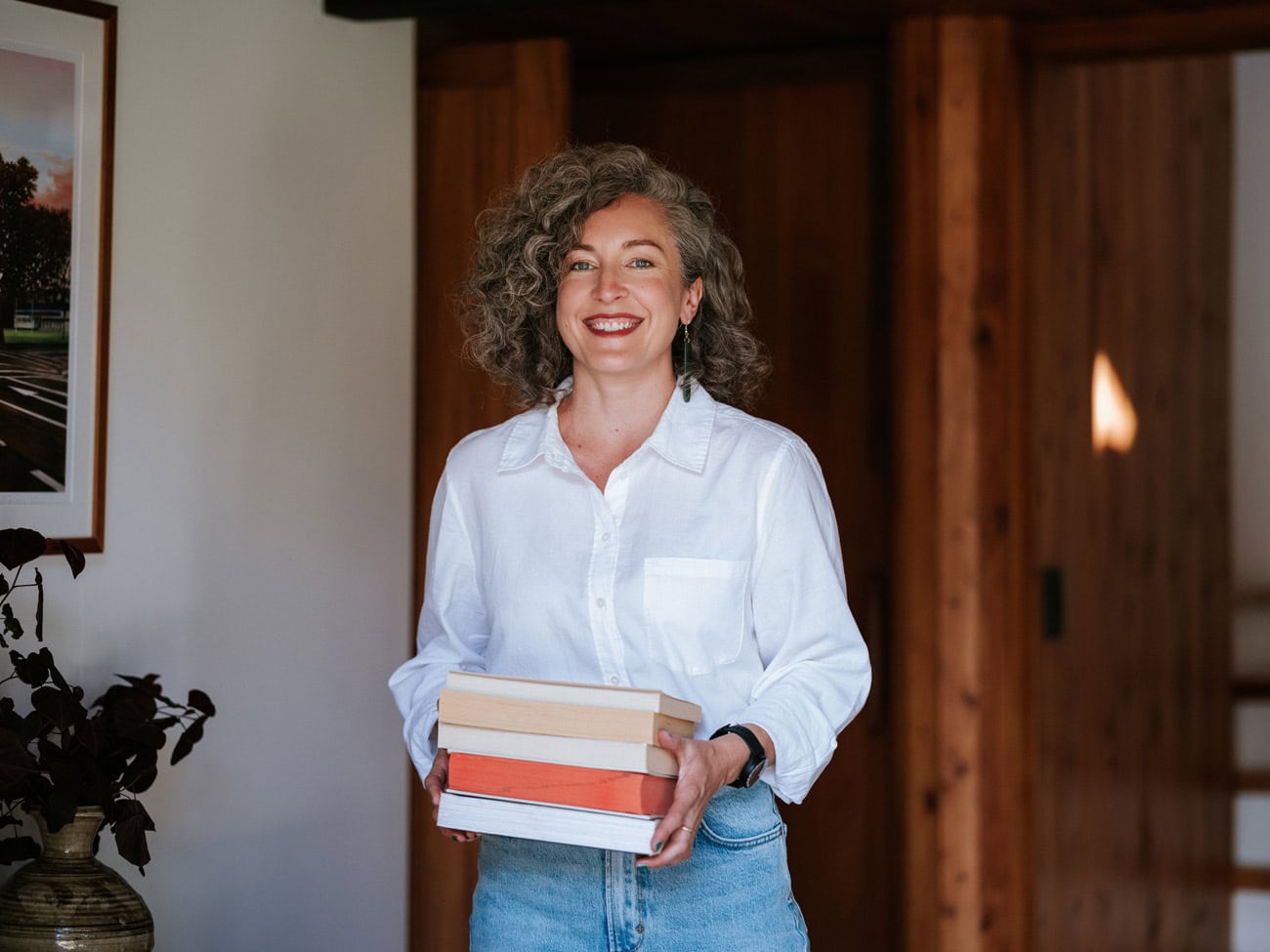 Photo by Florence Charvin Photography of A woman with curly grey hair, wearing a white shirt and jeans, stands indoors holding a stack of books and smiling at the camera.
