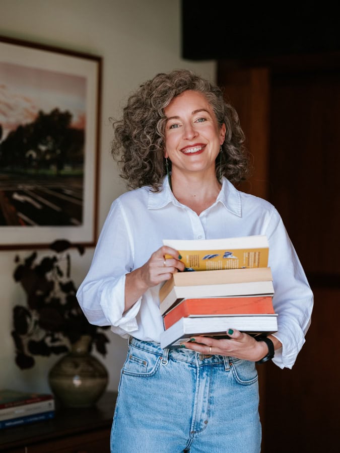 Photo by Florence Charvin Photography of A person with curly hair and a white shirt holds a stack of books and smiles whilst standing indoors in a room with framed art and a vase in the background.