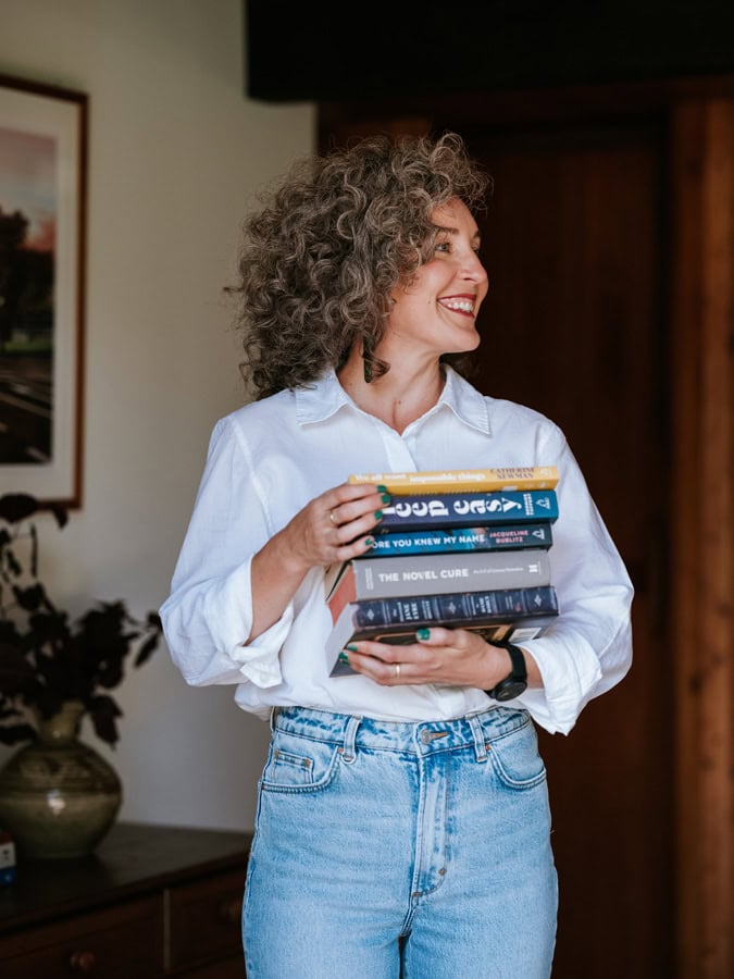 Photo by Florence Charvin Photography of A woman with curly hair, wearing a white shirt and jeans, is holding a stack of books and smiling whilst standing indoors.