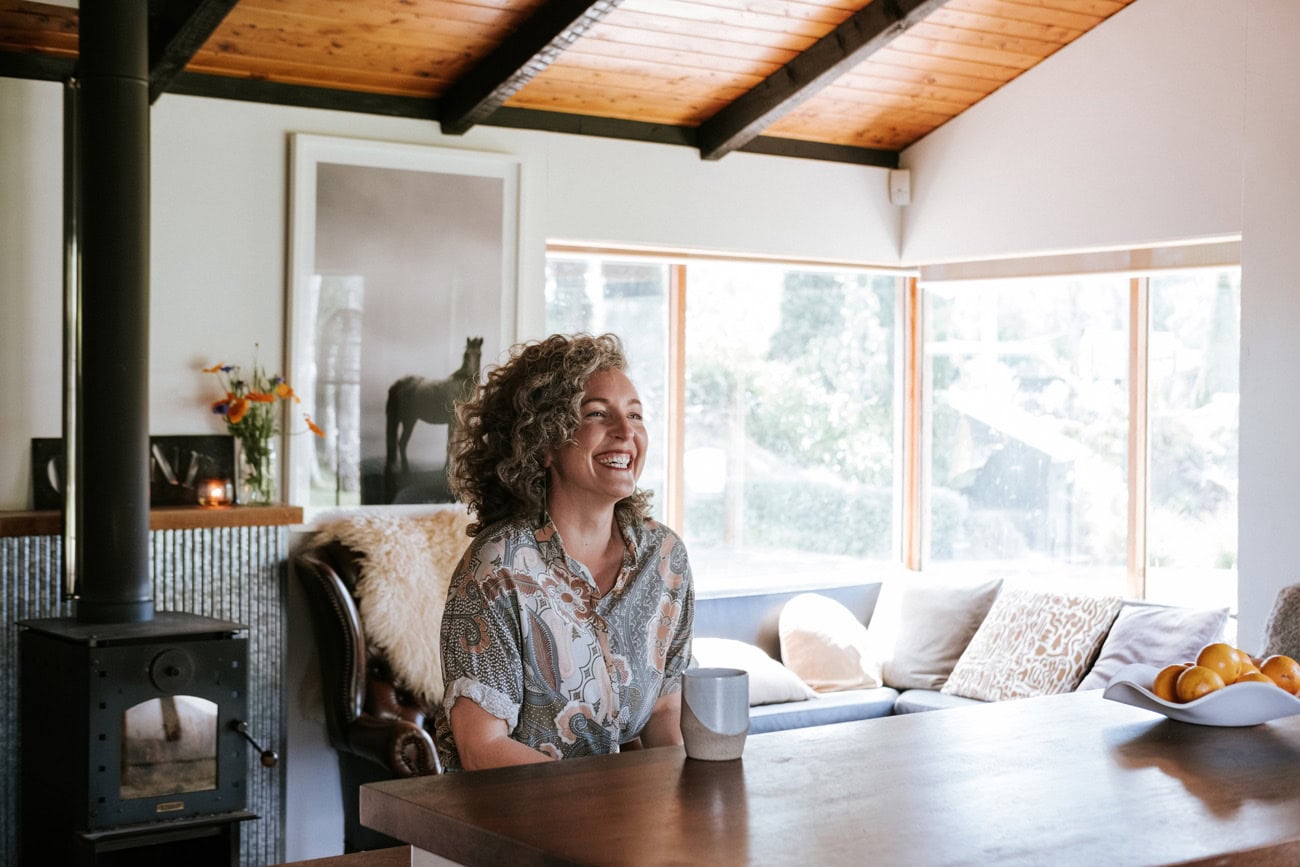 Photo by Florence Charvin Photography of A woman with curly hair sits at a kitchen worktop, smiling and holding a mug, with a wood-burning stove, sofa, and large windows in the background.