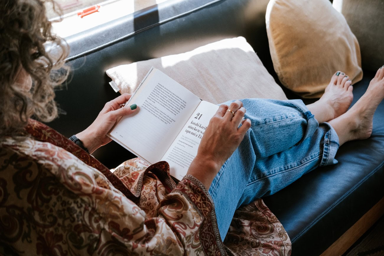 Photo by Florence Charvin Photography of A person with curly hair sits on a sofa with legs stretched out, reading an open book. They wear a patterned shirt and jeans, and a beige cushion is behind them.