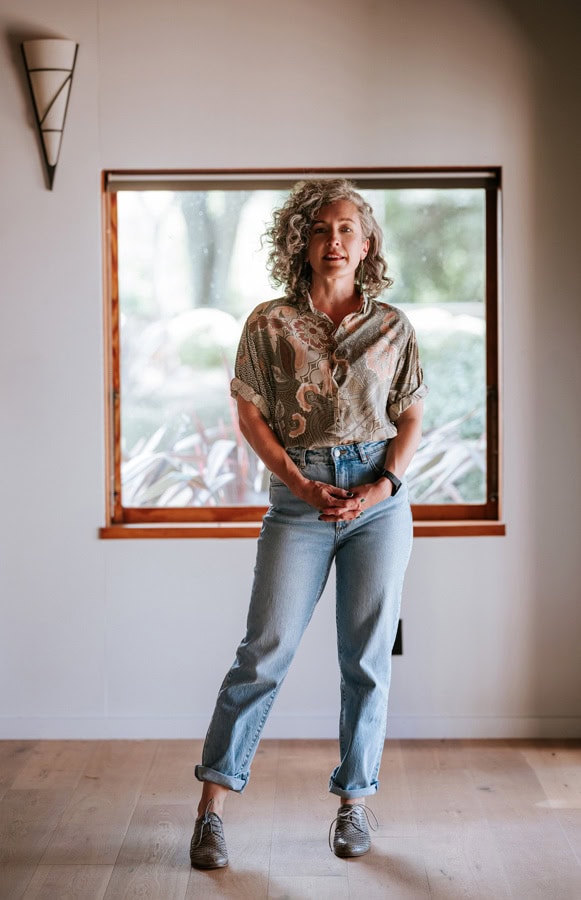 Photo by Florence Charvin Photography of A person with curly grey hair stands indoors in front of a window, wearing a patterned shirt, light blue jeans, and grey shoes.