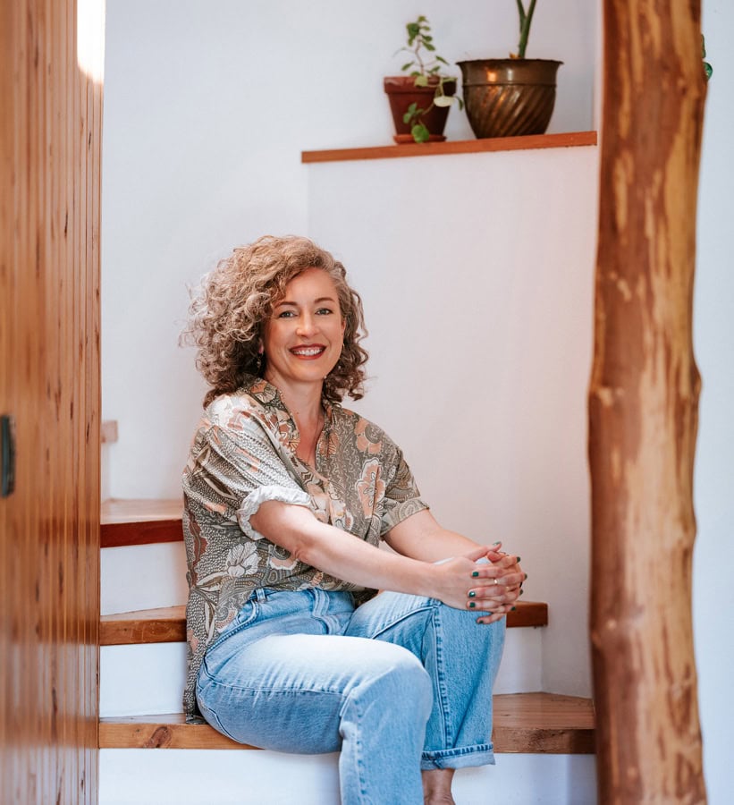 Photo by Florence Charvin Photography of A woman with curly hair sits on wooden stairs indoors, smiling, with potted plants on a shelf behind her.