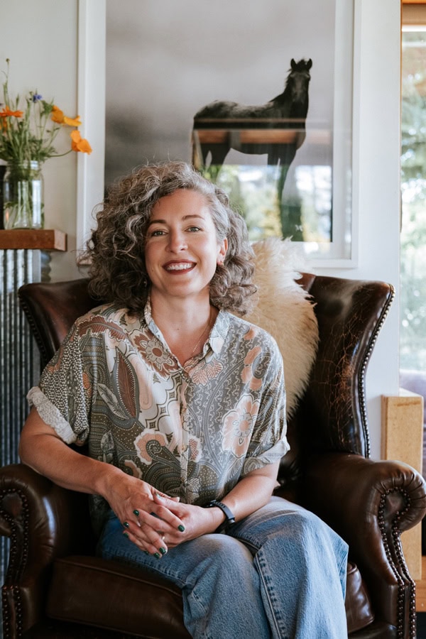 Photo by Florence Charvin Photography of A woman with curly hair sits smiling in a leather armchair, wearing a floral shirt and jeans. Behind her is a framed horse photograph and a vase of flowers on a shelf.