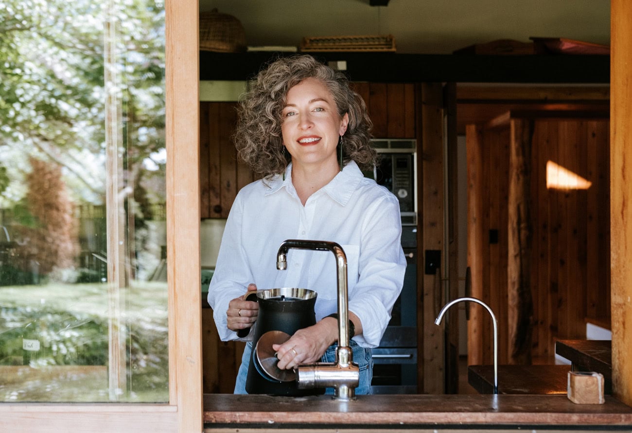 Photo by Florence Charvin Photography of A person with curly hair wearing a white shirt stands in a wooden kitchen by a sink, holding a jug and smiling at the camera.