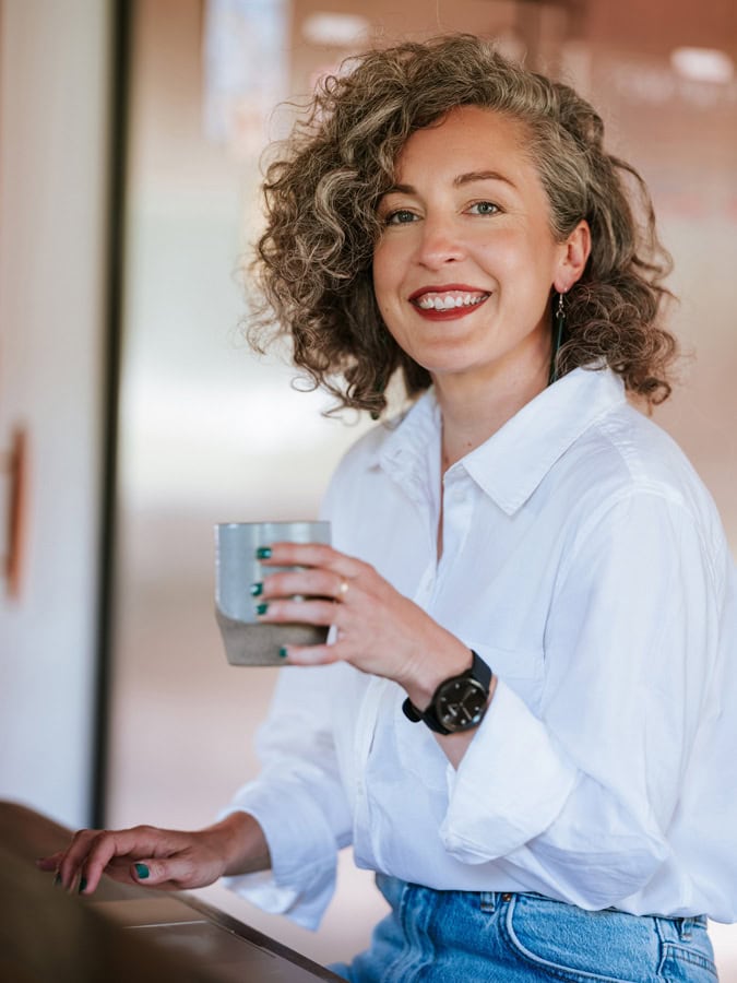 Photo by Florence Charvin Photography of A person with curly grey hair, wearing a white shirt and blue jeans, sits and smiles while holding a mug. They have green nail varnish and wear a black wristwatch.