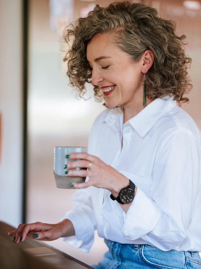 Photo by Florence Charvin Photography of Woman with curly hair wearing a white shirt and jeans, smiling whilst holding a mug and sitting indoors.