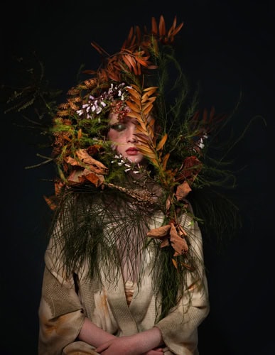 Photo by Florence Charvin Photography of A person wearing a beige dressing gown is partially obscured by an elaborate arrangement of dried leaves, flowers, and foliage against a dark background.