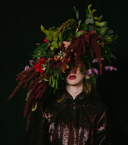 Photo by Florence Charvin Photography of A person wearing a shiny, dark outfit is partially obscured by an elaborate floral headpiece covering much of their face, set against a black background.
