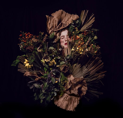 Photo by Florence Charvin Photography of A person’s face is partially visible among an elaborate arrangement of dried leaves, flowers, and textured brown material against a dark background.