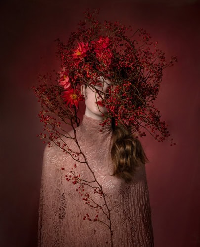 Photo by Florence Charvin Photography of A person wearing a lace garment stands against a reddish background with their face partially obscured by a large arrangement of red berries and flowers.