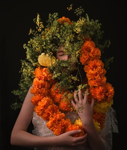 Photo by Florence Charvin Photography of A person partially obscured by a large arrangement of orange and yellow flowers and greenery against a dark background.