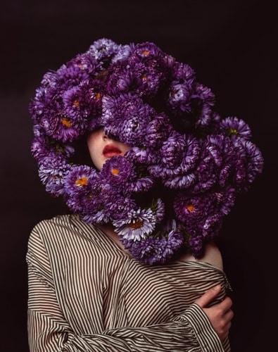 Photo by Florence Charvin Photography of A person wearing a striped shirt with their face obscured by a large arrangement of purple flowers against a dark background.
