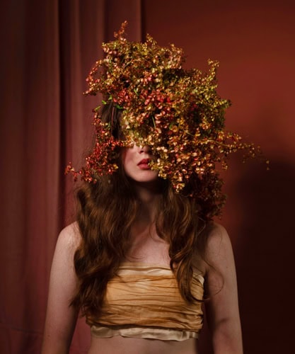 Photo by Florence Charvin Photography of A woman with long brown hair wears a beige bandeau top. Her face is partially obscured by a large bouquet of red and yellow flowers. The background is a reddish-brown curtain.