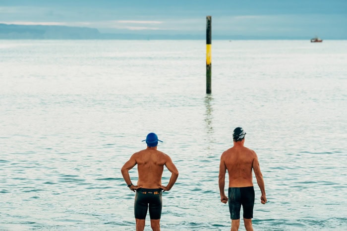 Photo by Florence Charvin Photography of Two men wearing swimming caps and swimming trunks stand at the edge of a calm sea, facing the water with a marker pole visible in the distance.