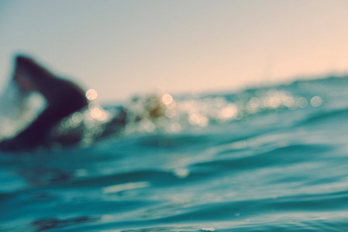 Photo by Florence Charvin Photography of Out-of-focus image of a person in dark clothing in the water, with sunlight reflecting off the surface and a blurred horizon in the background.