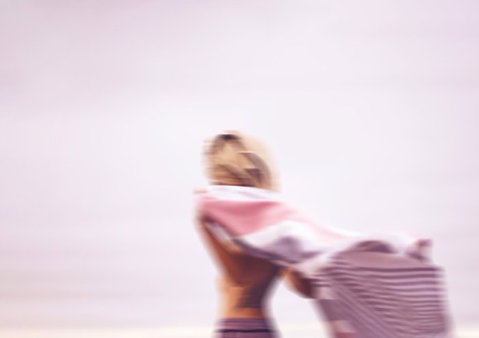 Photo by Florence Charvin Photography of A person with long hair stands near the shore, holding a striped towel or blanket. The image is out of focus and the background appears to be a beach or open water.