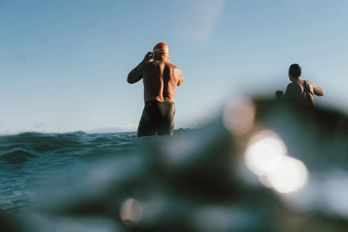 Photo by Florence Charvin Photography of Two people in swimming caps and goggles stand in the sea, partially submerged, preparing for a swim under a clear sky.