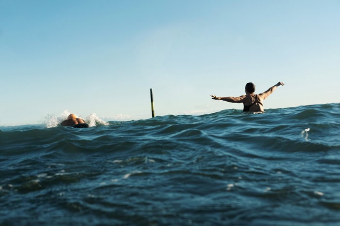 Photo by Florence Charvin Photography of Two people swim in the sea, one with arms outstretched facing away from the camera, the other swimming forwards; a pole is visible in the distance under a clear sky.