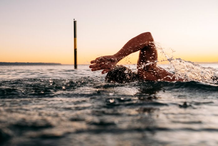 Photo by Florence Charvin Photography of Person swimming front crawl in open water at sunset, with water splashing and a distant pole visible in the background.