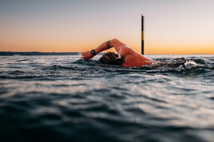 Photo by Florence Charvin Photography of A person swimming front crawl in open water at sunset, with their head down and one arm extended forward above the surface.