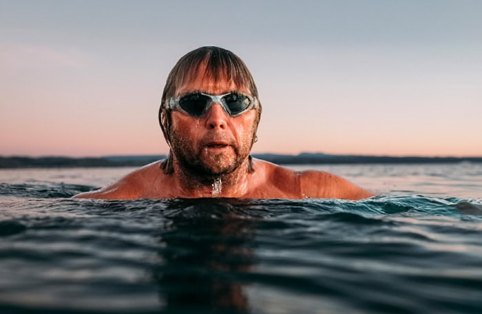 Photo by Florence Charvin Photography of A man wearing swimming goggles is partially submerged in open water, with a sunset sky and distant land visible in the background.