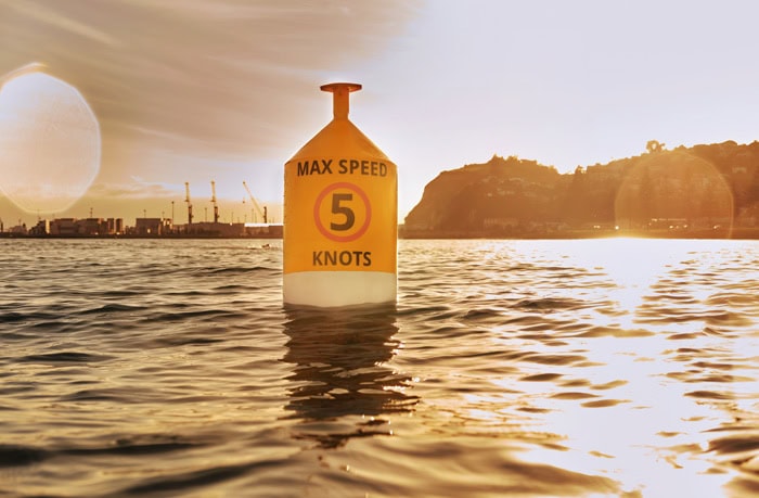 Photo by Florence Charvin Photography of A yellow buoy in the water displays a "Max Speed 5 Knots" sign, with a shoreline and industrial cranes visible in the background at sundown.