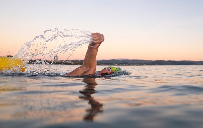 Photo by Florence Charvin Photography of A swimmer wearing a yellow cap and goggles performs a front crawl stroke in open water during sunset or sunrise.