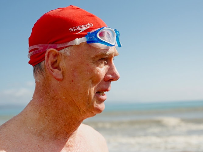 Photo by Florence Charvin Photography of Older man wearing a red swimming cap and blue goggles stands by the sea, wet from swimming, with ocean waves and sky in the background.