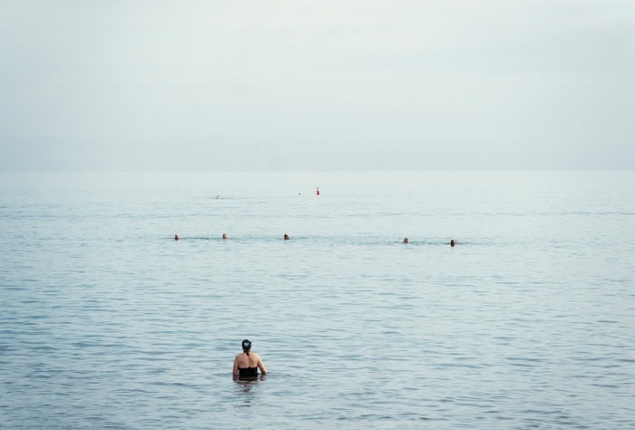 Photo by Florence Charvin Photography of A person in swimwear stands in shallow water facing the open sea, with several buoys floating in the distance under an overcast sky.