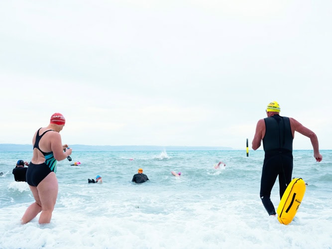 Photo by Florence Charvin Photography of Several swimmers wearing wetsuits and swim caps enter the sea, with a few already swimming further out in the water under an overcast sky.